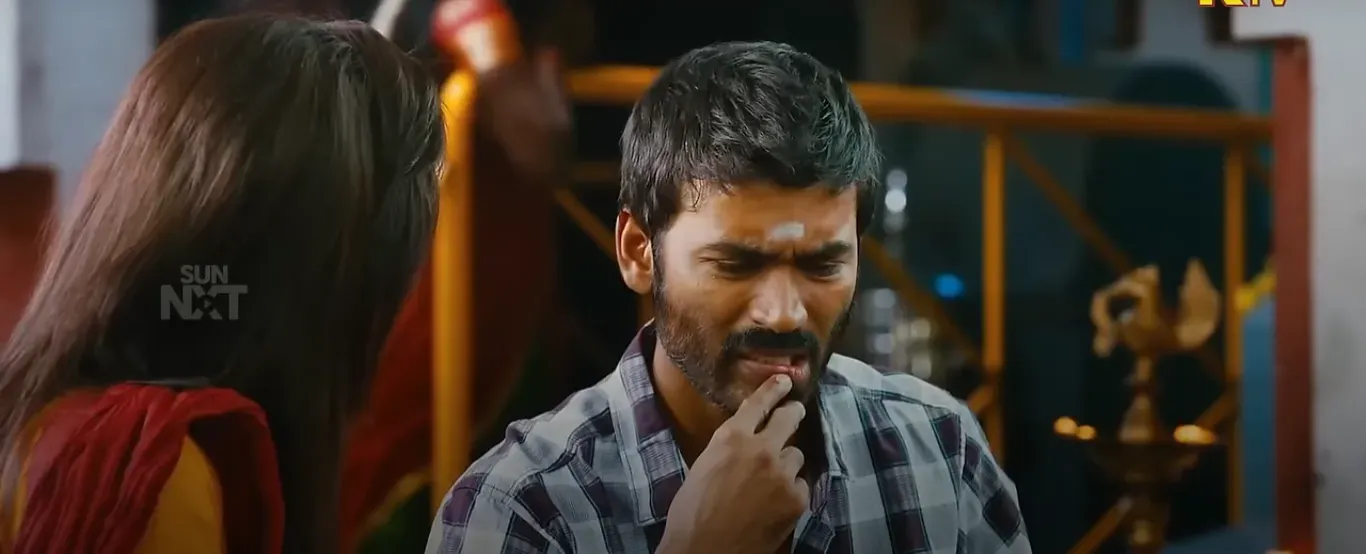 Dhanush with a confused expression, finger near his mouth, looking lost in thought.