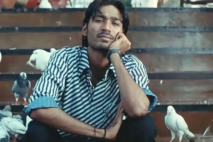 Dhanush resting his cheek against his hand while sitting amidst white pigeons on steps