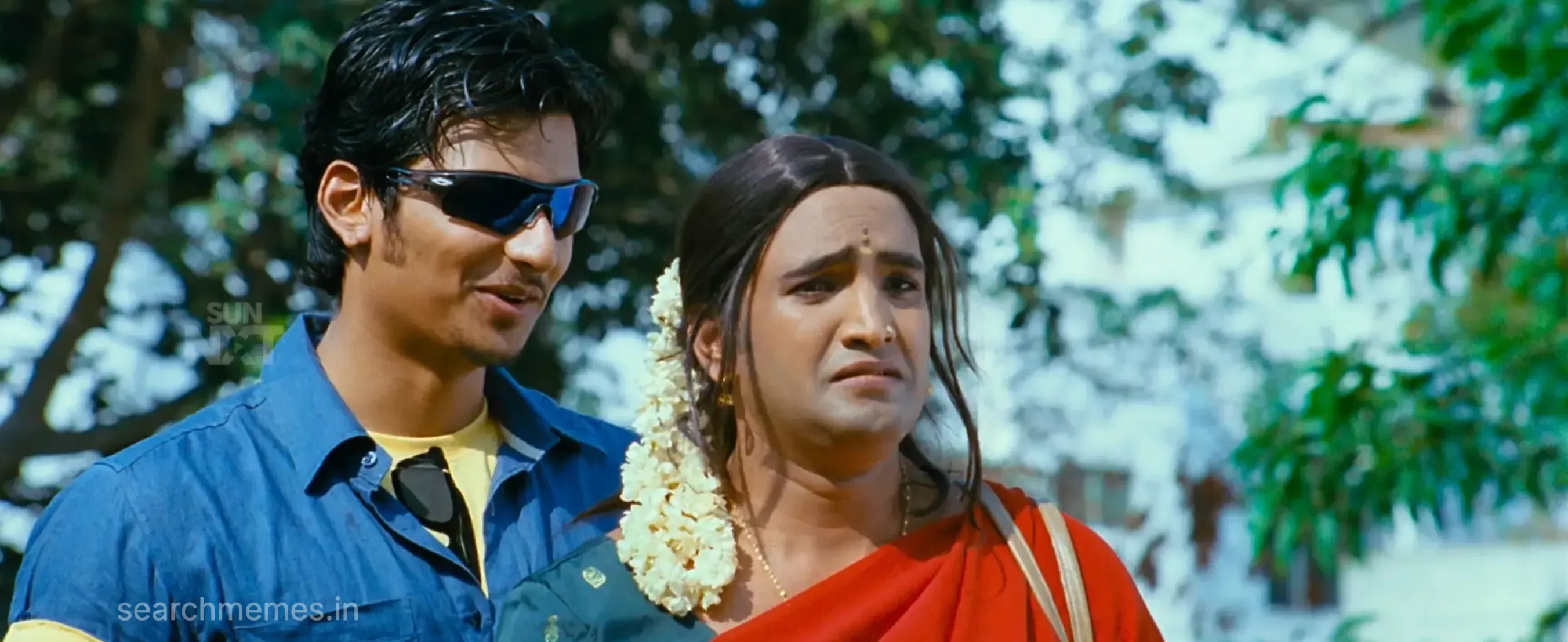 Jiiva in sunglasses smiling next to Santhanam dressed in a saree and jasmine flowers