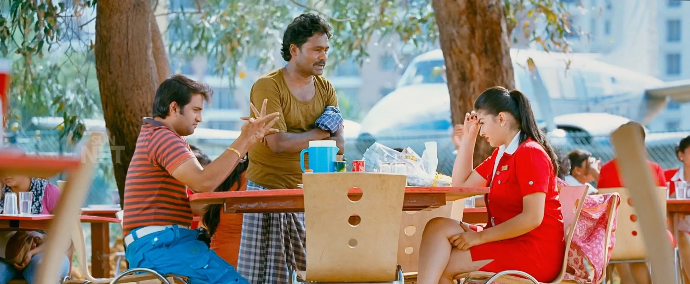 Santhanam gesturing animatedly at an outdoor cafe table with Hansika in OK OK