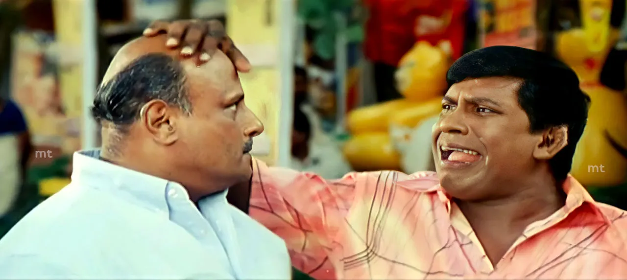 Vadivelu grinning widely with his hand placed on another man's bald head in Muruga