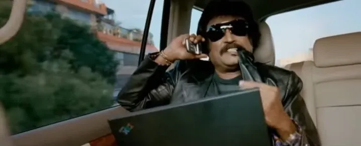 Rajinikanth in sunglasses holding a phone in a car with a laptop