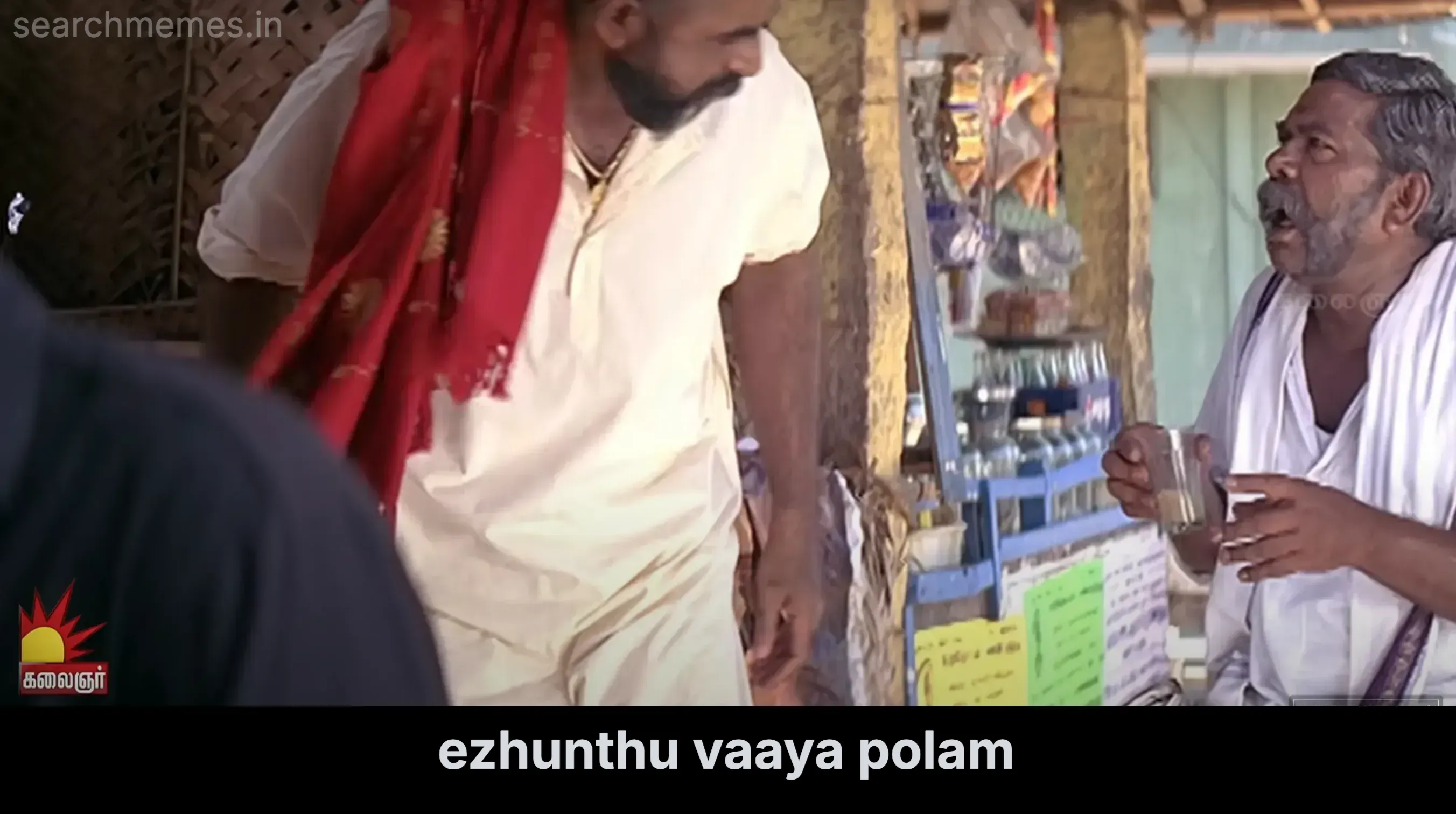 Karthi standing up with a red cloth while Saravanan sits holding a glass of tea at a stall