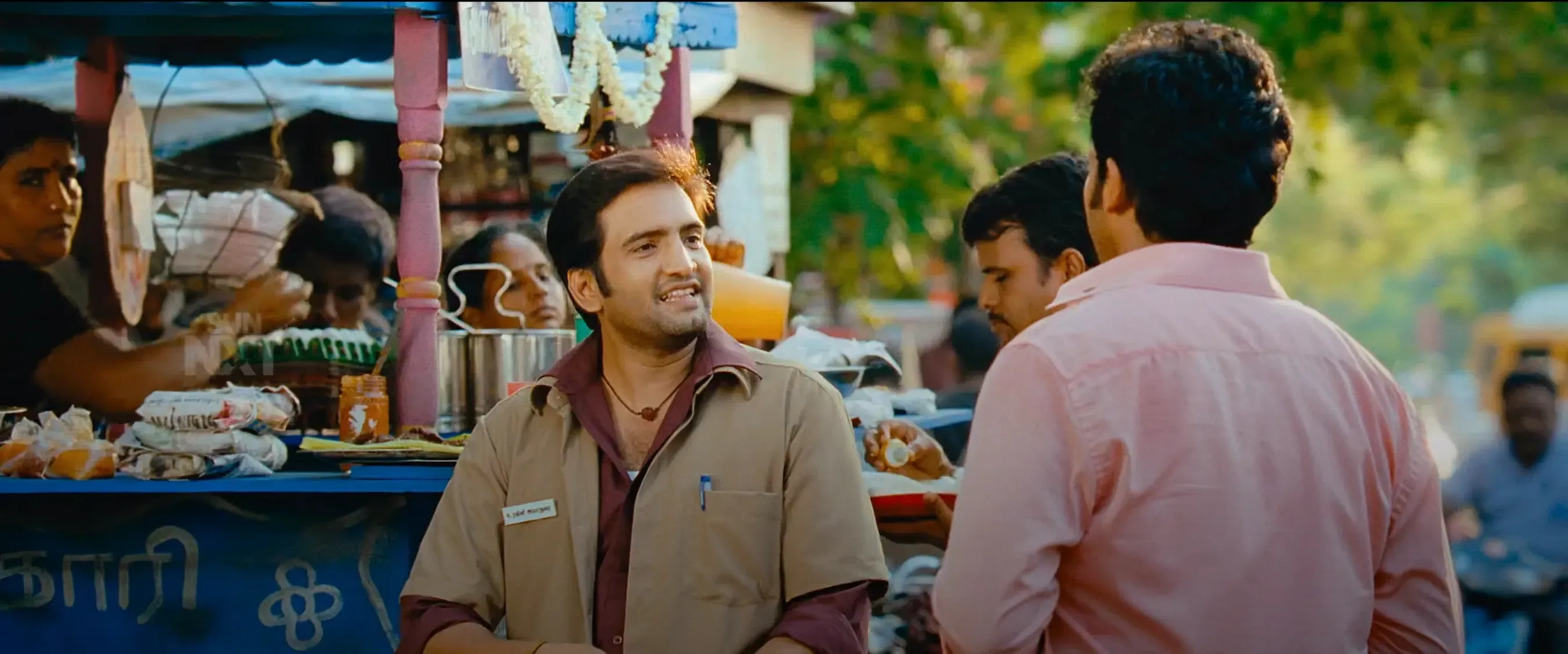 Santhanam in khaki uniform smiling and talking at a tea stall in Saguni