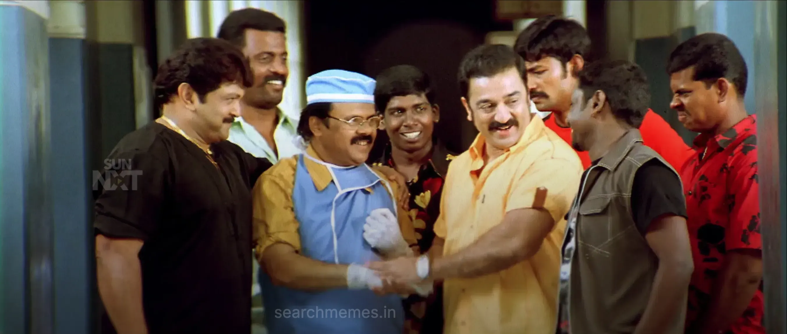 Kamal Haasan smiling and shaking hands in a yellow shirt surrounded by friends