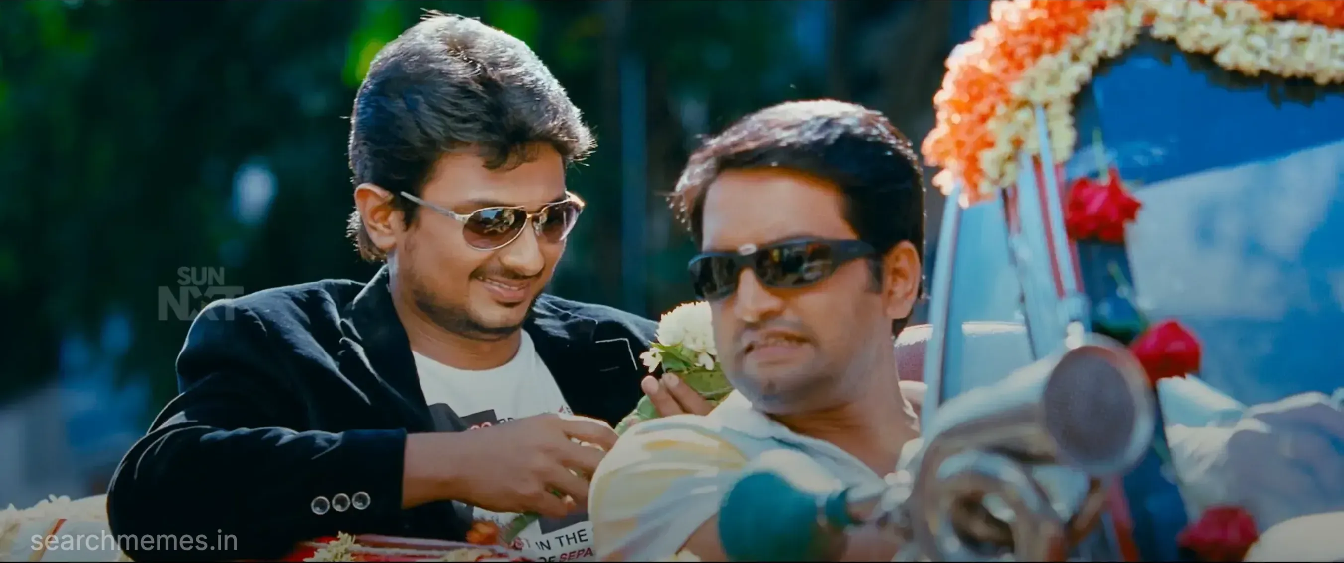 Udhayanidhi smiles as he hands flowers to an annoyed Santhanam wearing sunglasses in a car