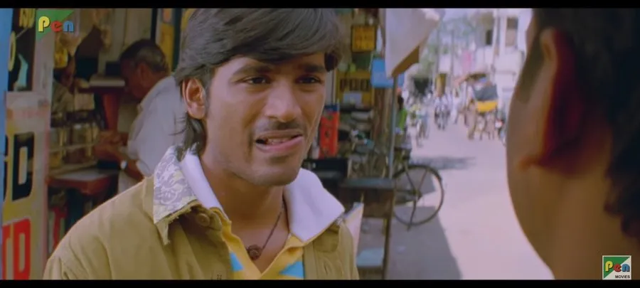 Dhanush talking sideways with a quizzical smirk on a busy street corner