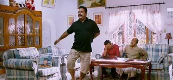 Thambi Ramaiah standing and posing awkwardly in a living room while two men sit on the couch