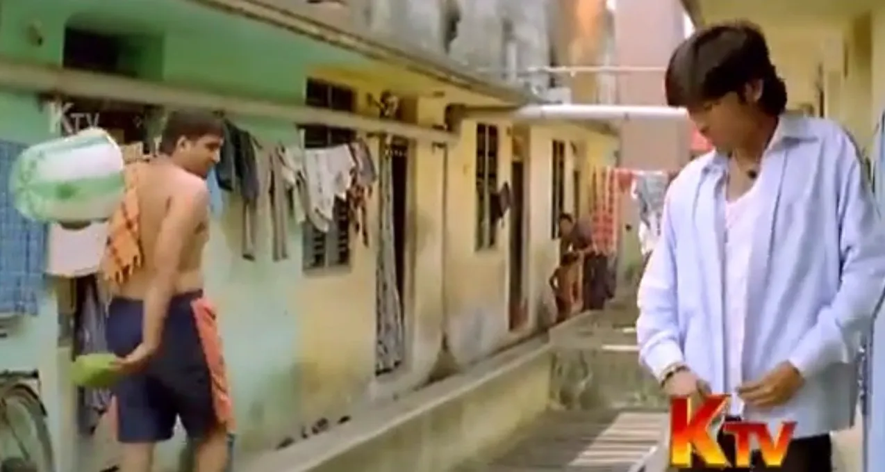Dhanush buttoning his shirt while Santhanam in shorts holds a water jug in a house corridor
