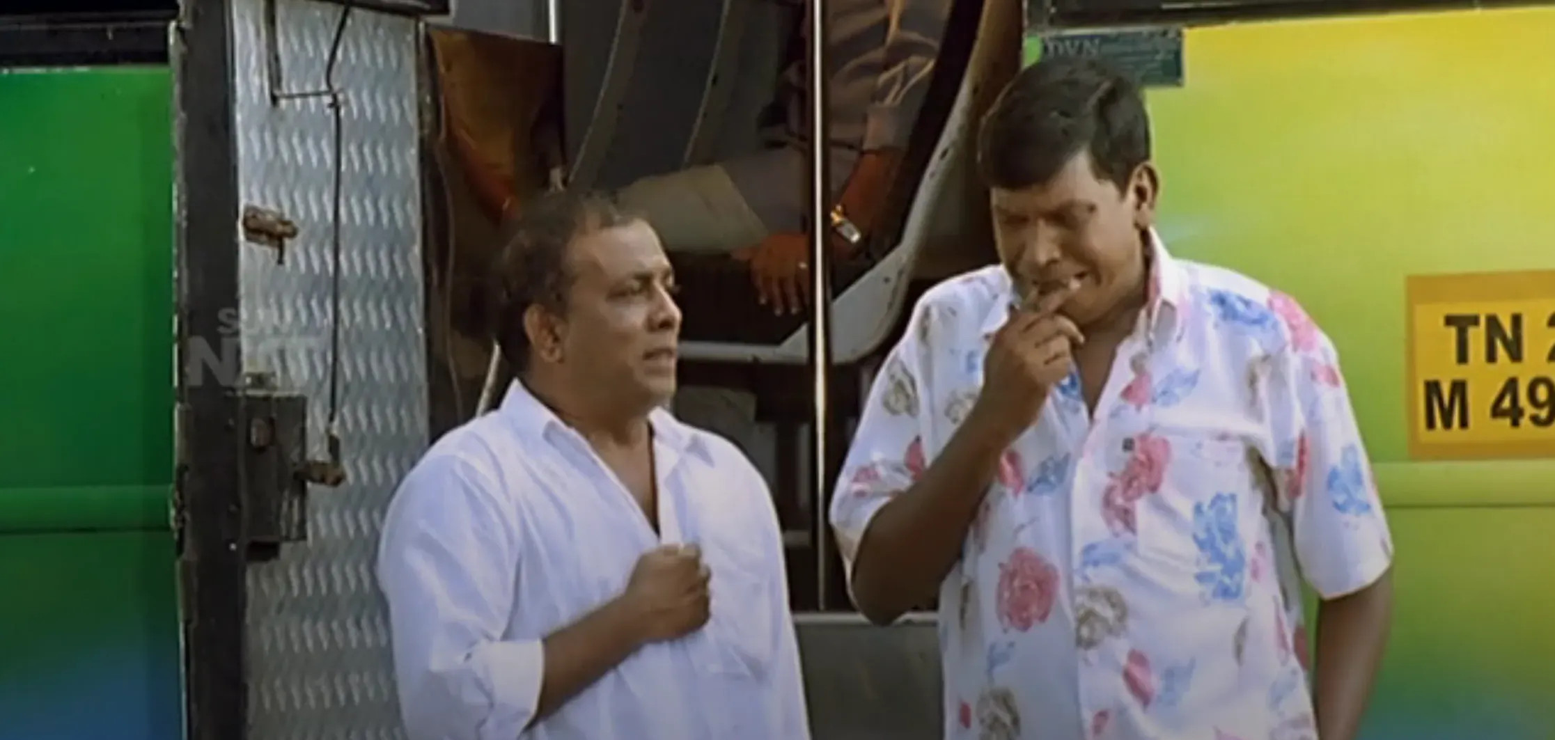 Singamuthu holding his chest and Vadivelu biting his finger in deep thought outside a green bus