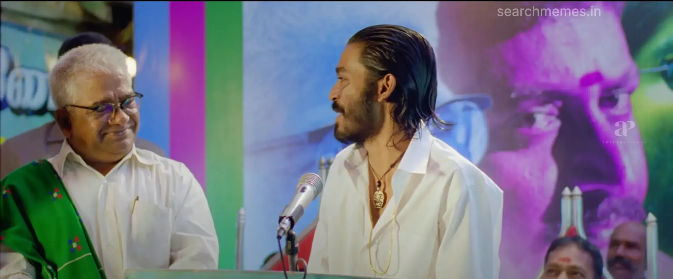 Dhanush at a podium speaking into a microphone with an older man beside him in Pudhupettai.