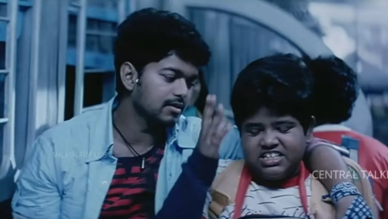 Vijay in denim jacket raising hand beside a chubby boy in a bus