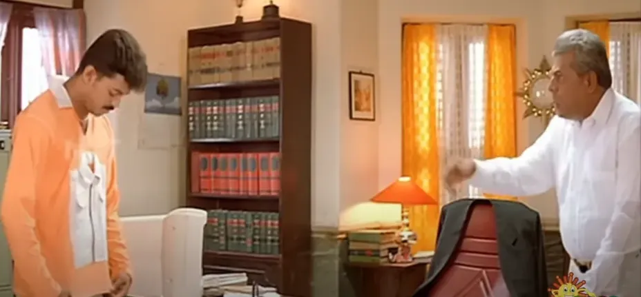 Delhi Ganesh gesturing sternly at Vijay standing politely beside office table