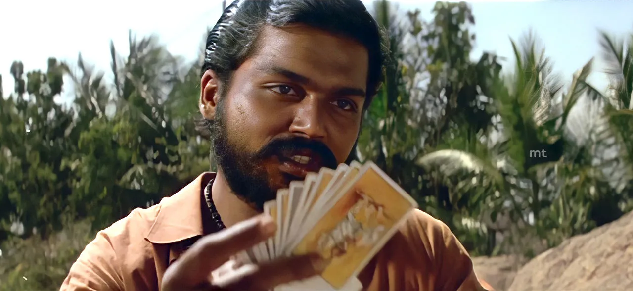 Karthi in rural attire smiling victoriously over a card game outdoors